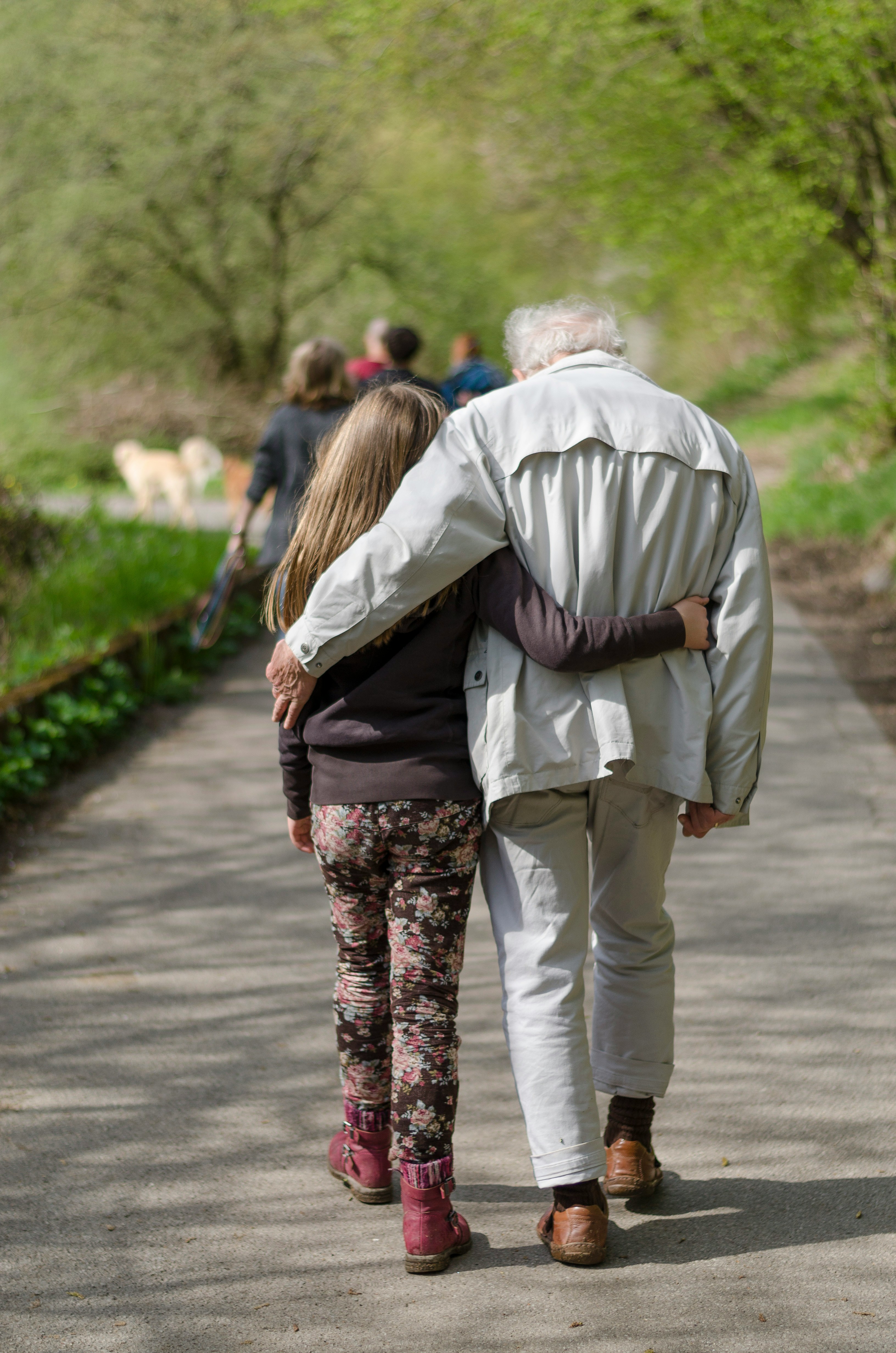A young woman and an older person walking arm in arm down a tree-lined path, seen from behind, on a sunny spring day
